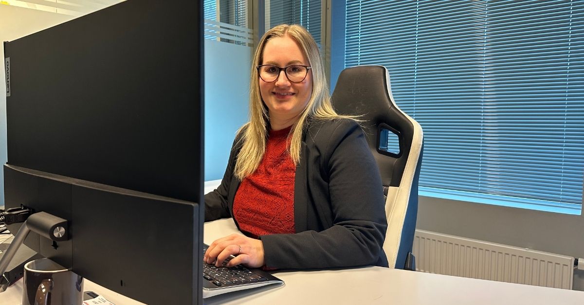 Kvinne sitter foran pc-skjerm på et kontor. Smiler til kamera og med hendene på tastaturet. 