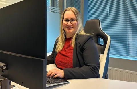 17 tips til bedre wifi hjemme Kvinne sitter foran pc-skjerm på et kontor. Smiler til kamera og med hendene på tastaturet.